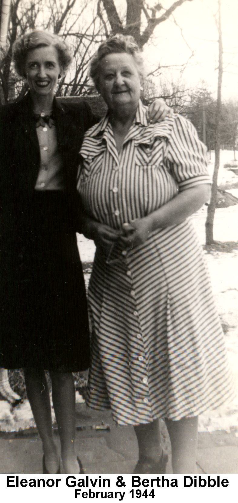 Black and white photo of Eleanor Galvin with short hair parted on the left, wearing a black knee-length skirt and blazer with a lighter-colored blouse standing with her left arm around the shoulders of Bertha Dibble, with curly grey hair and wearing a pin-striped calf-length dress, on a snow-covered winter day in front of bare trees.